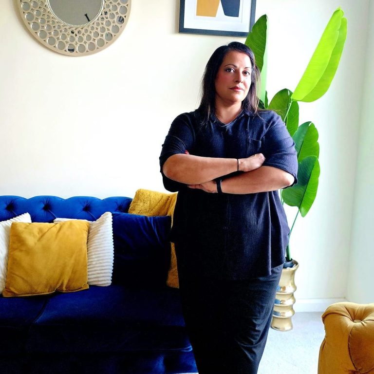 Sara Female therapist and counsellor stands with arms crossed in a calming, stylish room with a blue sofa and decorative cushions.