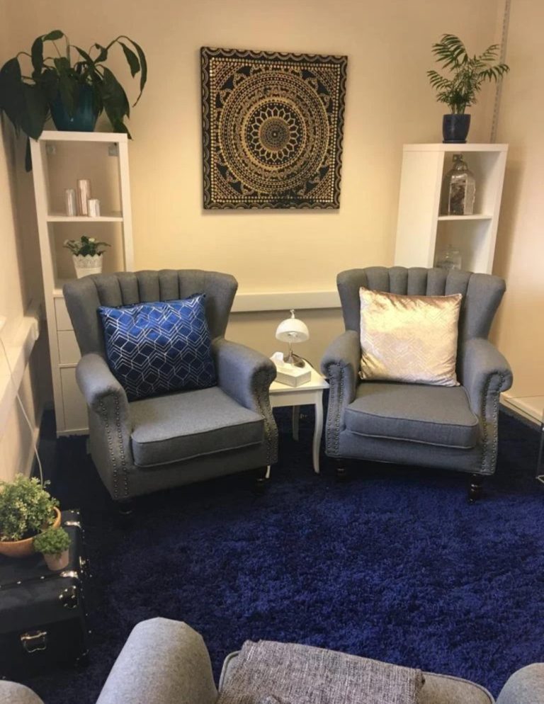 Therapy session Two comfortable armchairs with blue cushions, a side table, and a patterned wall hanging in a therapist room.