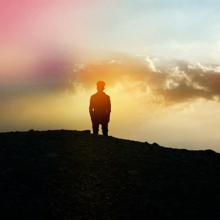 Hopeful, reflective, silhouetted figure standing on a hill against a colourful sunset sky.