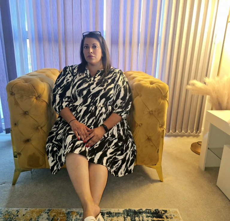 Sara Female therapist in a black and white dress sits on a comforting yellow armchair in a bright room.